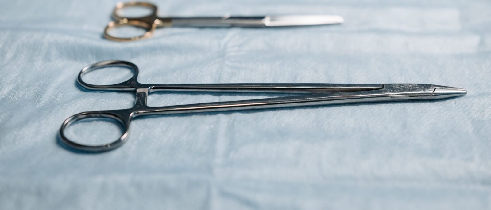 Close-up de instrumentos cirúrgicos incluindo porta-agulha em pano azul em ambiente médico