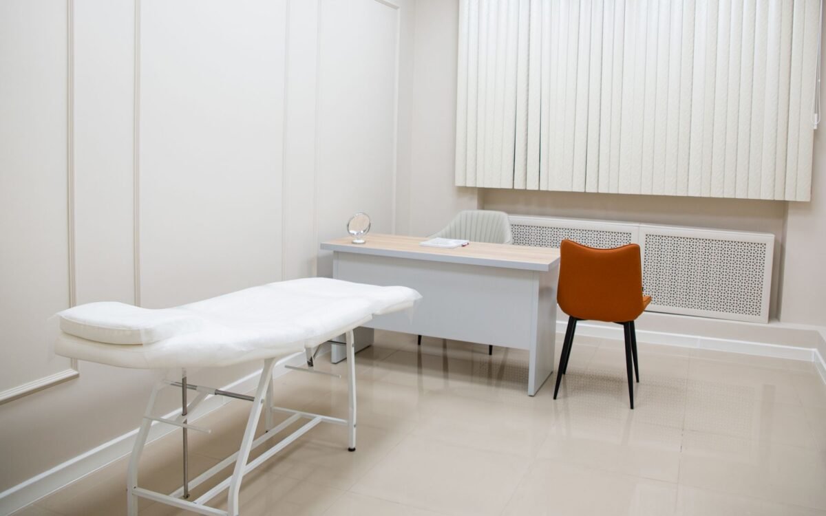 Consultório médico minimalista com escrivaninha, cadeiras e maca de exame em ambiente estéril e neutro